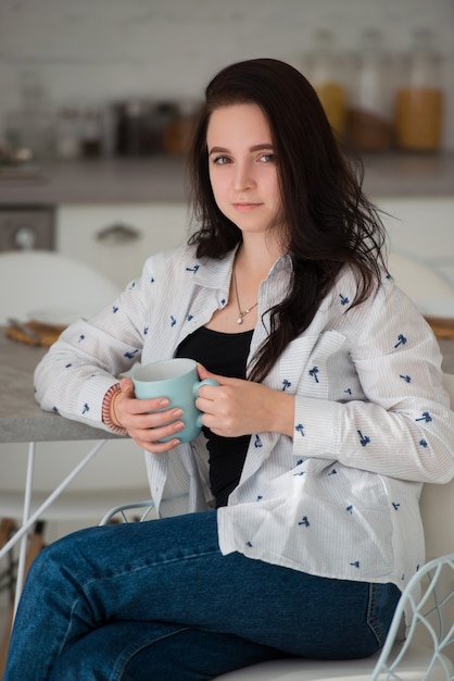 La giovane studentessa bruna si siede su una sedia al mattino in cucina con una tazza di caffè