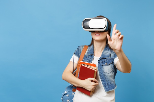 La giovane studentessa in occhiali per realtà virtuale tiene i libri toccando qualcosa come premere il pulsante, indicando lo schermo virtuale galleggiante isolato su sfondo blu. Istruzione nel college universitario scolastico.