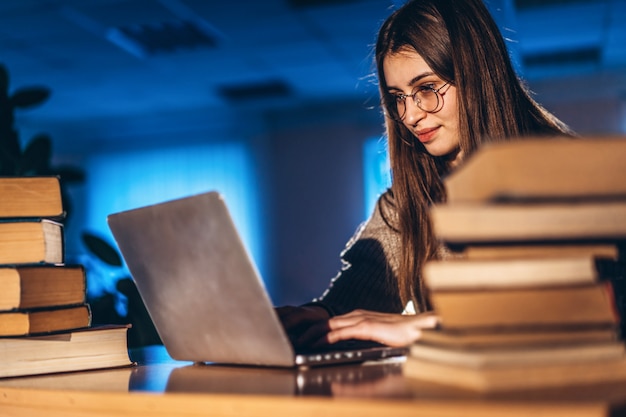 La giovane studentessa la sera si siede a un tavolo della biblioteca con una pila di libri e lavora su un computer portatile. Preparazione per l'esame