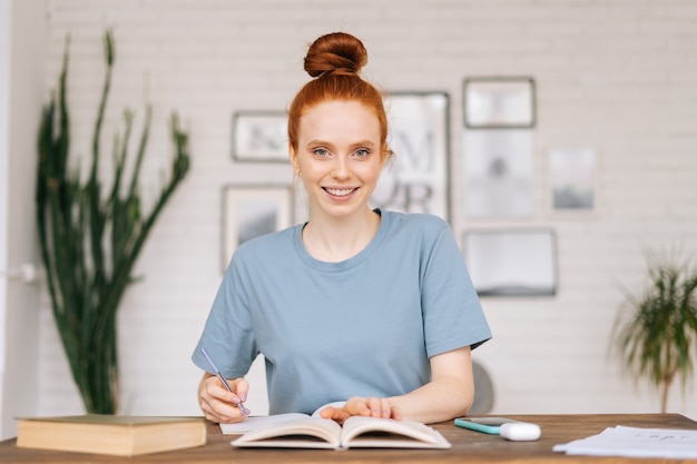 La giovane studentessa sorridente felice della testarossa sta facendo un profilo dal libro di testo nella cartella di lavoro