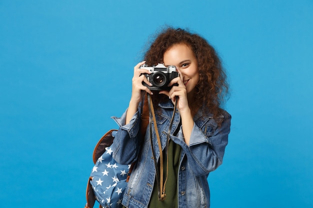 La giovane studentessa teenager della ragazza afroamericana in vestiti del denim tiene la macchina fotografica isolata sulla parete blu