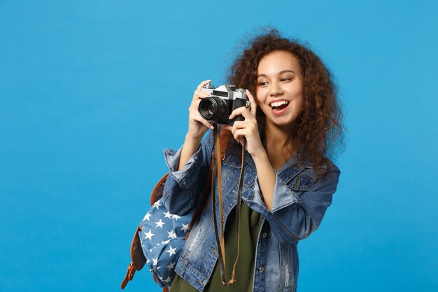 La giovane studentessa teenager della ragazza afroamericana in vestiti del denim tiene la macchina fotografica isolata sulla parete blu