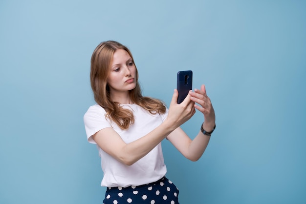 La giovane vlogger con la faccia sorpresa si fotografa al telefono mentre trasmette su uno sfondo blu con una t-shirt bianca. Bella ragazza caucasica. Selfie concept story e blog