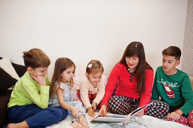 La grande famiglia felice si diverte insieme in camera da letto. Grande concetto di mattina in famiglia. La madre con quattro bambini indossa un pigiama e legge un libro a letto a casa.