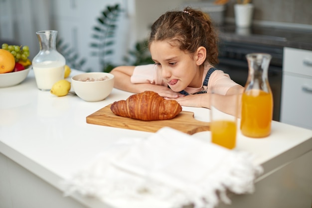 La graziosa bambina con i capelli ricci stava guardando un delizioso croissant al tavolo della cucina.