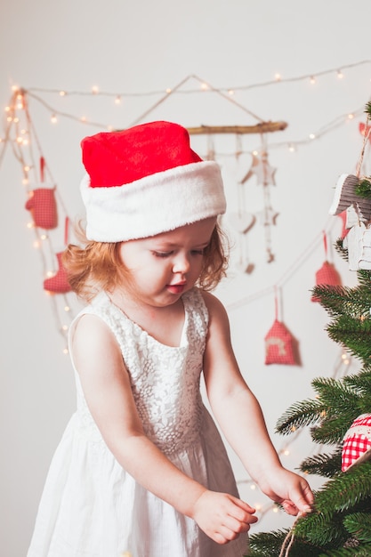 La graziosa bambina con un berretto di Babbo Natale sta decorando l'albero di Natale