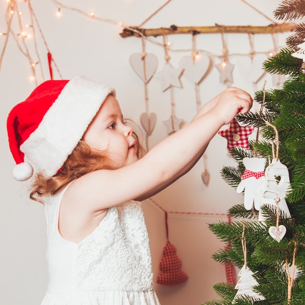 La graziosa bambina con un berretto di Babbo Natale sta decorando l'albero di Natale