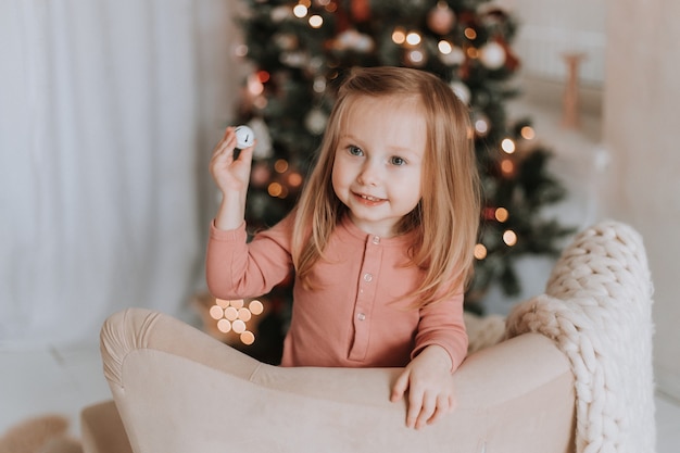 la graziosa ragazzina bionda è inginocchiata su una sedia sullo sfondo di un albero di Natale