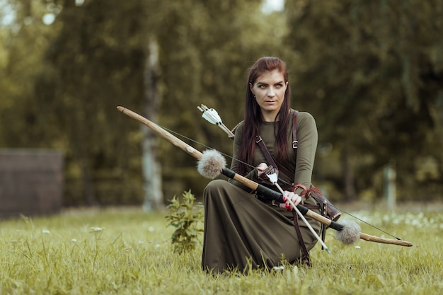 La guerriera medievale con un arco siede in una radura, a caccia in una foresta verde