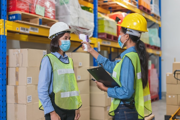 La lavoratrice asiatica indossa la maschera per il viso nel giubbotto di sicurezza utilizzando la scansione a infrarossi del termometro per controllare la temperatura corporea con il collega prima di lavorare nella fabbrica del magazzino durante la pandemia di coronavirus