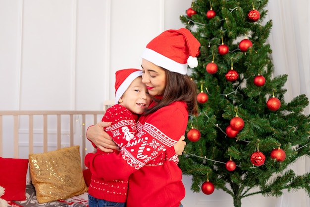 La madre abbraccia un bambino in maglioni rossi vicino all'albero di Natale