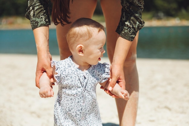 La madre aiuta la ragazza a muovere i primi passi sulla spiaggia Adorabile camminare insieme sulla sabbia in una giornata estiva Primo piano