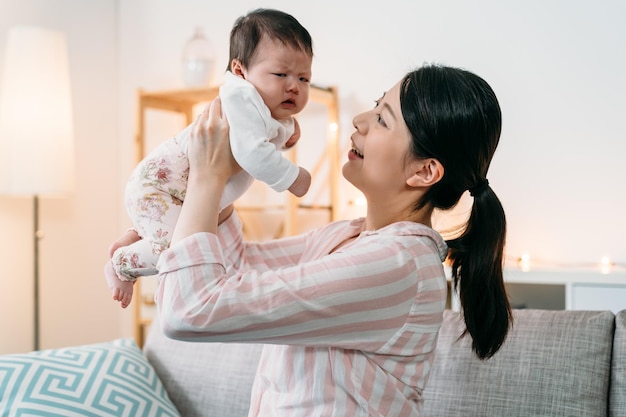 la madre asiatica affettuosa del ritratto laterale sta sollevando il suo bambino accigliato sconvolto con un sorriso mentre cerca di renderlo felice con un dolce comfort in un accogliente interno di casa.