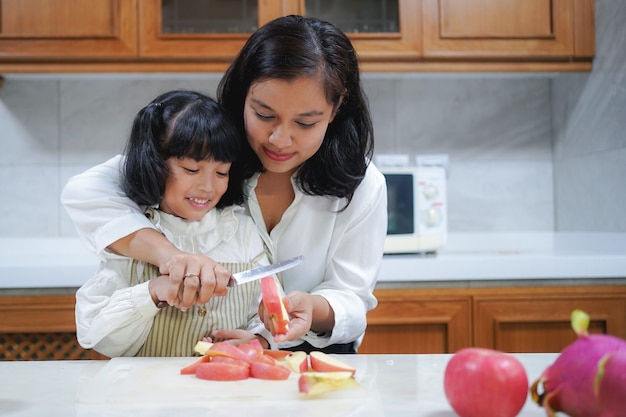 La madre asiatica sta insegnando alla sua piccola figlia a tagliare la mela in cucina a casa.