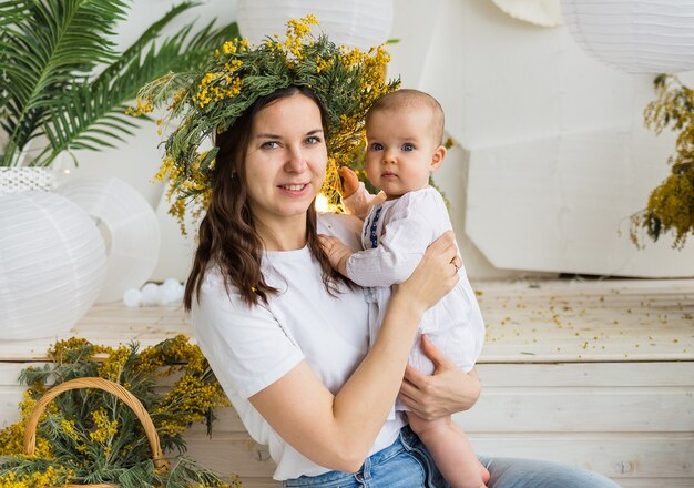 La madre con una ghirlanda di mimosa tiene in braccio un bambino in un abito bianco su uno sfondo di mazzi di mimosa