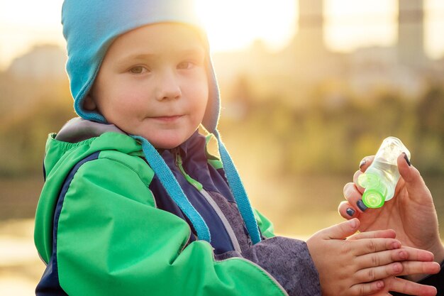 La madre con una giacca calda e un cappello lavorato a maglia applica un gel antisettico gel antibatterico bambino che cammina nel parco autunnale donna bionda elegante con un bambino sullo sfondo del lago