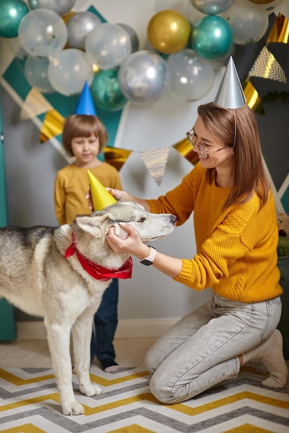 La madre e il bambino della famiglia di compleanno dell'animale domestico del cane mettono un cappello festivo sulla testa del cane congratulandosi per il compleanno