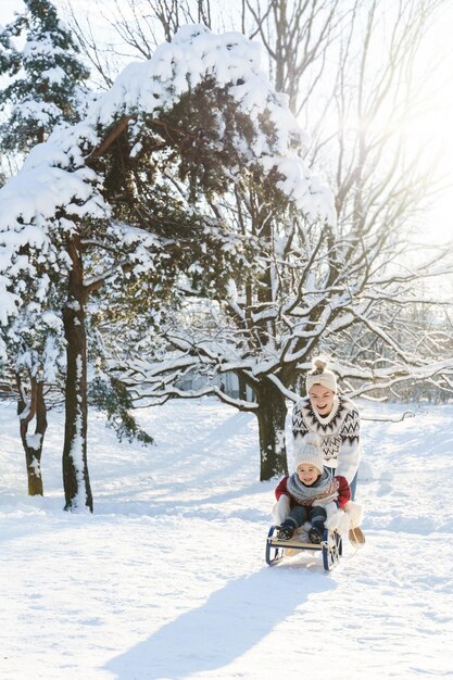 La madre e il suo simpatico figlioletto che indossano maglioni caldi si divertono su una collina per lo slittino durante la soleggiata giornata invernale