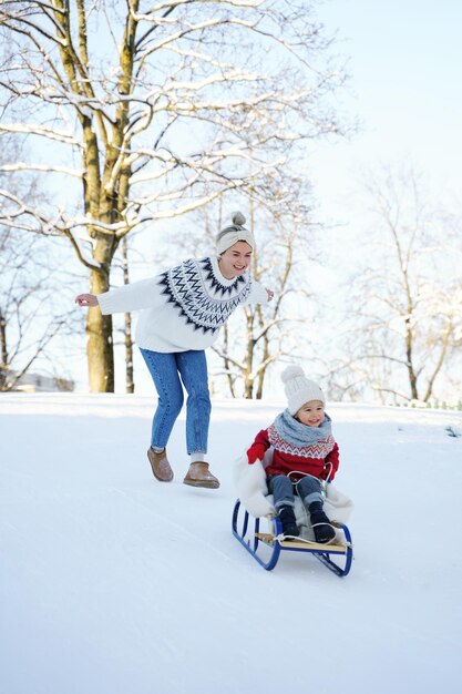 La madre e il suo simpatico figlioletto che indossano maglioni caldi si divertono su una collina per lo slittino durante la soleggiata giornata invernale