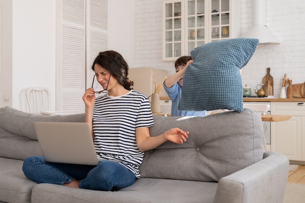 La madre esausta seduta sul divano a casa lavora sul laptop, il bambino distrae, attacca la mamma con il cuscino