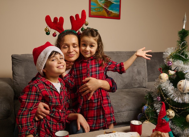 La madre felice con il cerchio di corna abbraccia i suoi adorabili bambini vestiti con abiti scozzesi rossi e verdi e un cappello da Babbo Natale sulla testa di suo figlio, celebrando la festa di Natale a casa nella cerchia familiare