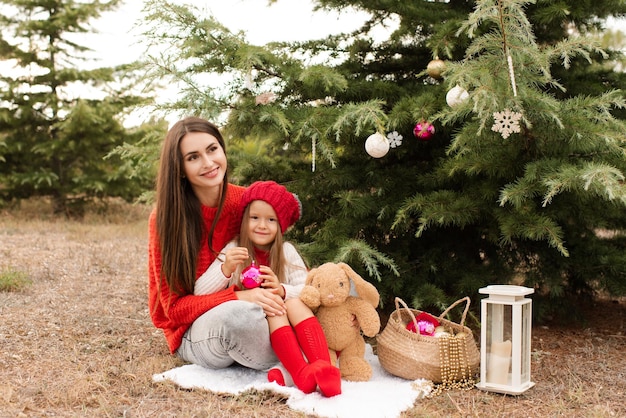 La madre felice con la bambina indossa maglioni a maglia rossi decorano l'albero di Natale all'aperto