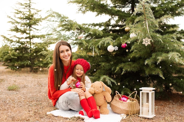 La madre felice con la bambina indossa maglioni a maglia rossi decorano l'albero di Natale all'aperto