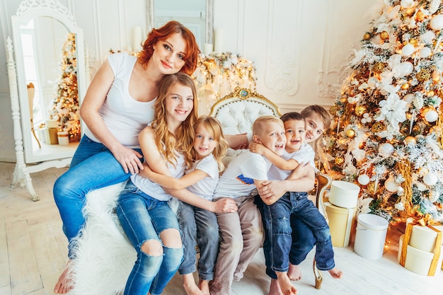 La madre felice della famiglia e cinque bambini si rilassano giocando vicino all'albero di Natale alla vigilia di Natale a casa. Figli della mamma figli nella stanza luminosa con decorazione invernale. Natale Capodanno per la celebrazione