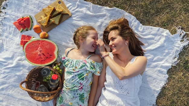 La madre felice tocca il naso della piccola figlia sdraiata su una coltre bianca con deliziosi frutti preparati per il picnic nella soleggiata giornata estiva vista dall'alto