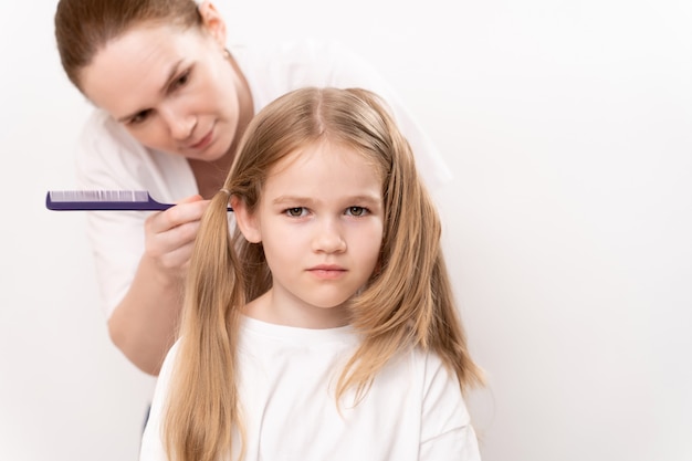 La madre pettina e fa i capelli di una bambina su uno sfondo bianco. La mamma è una parrucchiera. risparmiare denaro in un salone di bellezza. shampoo e cosmetici per i capelli dei bambini.