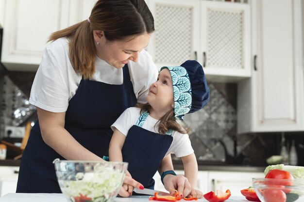 La madre premurosa insegna alla sua piccola figlia come preparare un'insalata in cucina, una giovane madre e un'affascinante dolce bambina si guardano e sorridono.