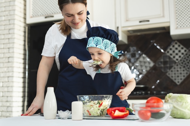 la madre premurosa insegna alla sua piccola figlia come preparare un'insalata in cucina, una giovane madre e un'affascinante dolce bambina vestire l'insalata, salarla e assaggiarla.