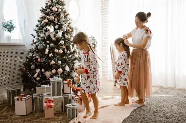 La madre premurosa intreccia la treccia della sua piccola figlia mentre la seconda figlia decora l'albero di Capodanno nella stanza luminosa e accogliente.