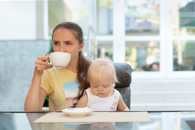 La mamma arrabbiata tiene il bambino in braccio e beve il caffè. Madre e mancanza di tempo.