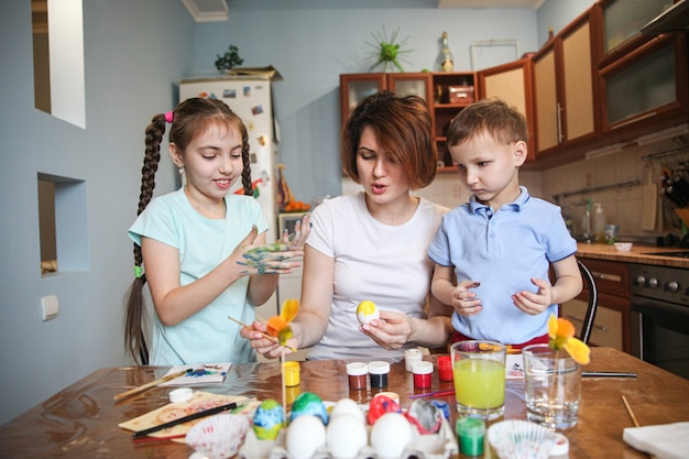 La mamma con due bambini decora le uova di Pasqua seduti a tavola a casa in cucina