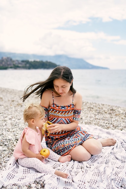 La mamma dà da mangiare alla bambina una banana mentre è seduta sulla spiaggia