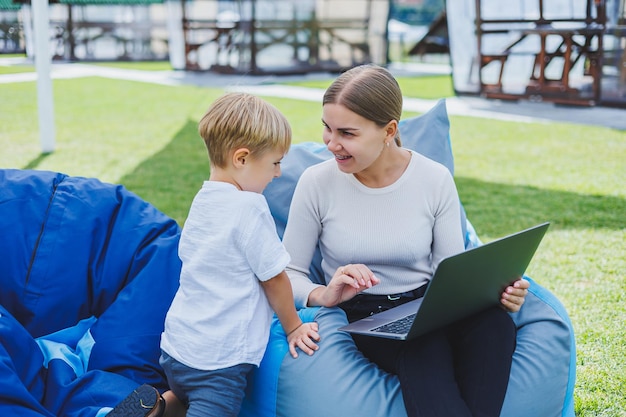 La mamma è in congedo di maternità Affascinante giovane donna con figlio piccolo che lavora all'aperto nel parco estivo con il laptop mentre è seduto su una sedia sull'erba verde