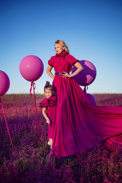 La mamma e la figlia di famiglia in abiti rosa sono in piedi in un campo con fiori e grandi palloncini al tramonto in estate contro il cielo blu