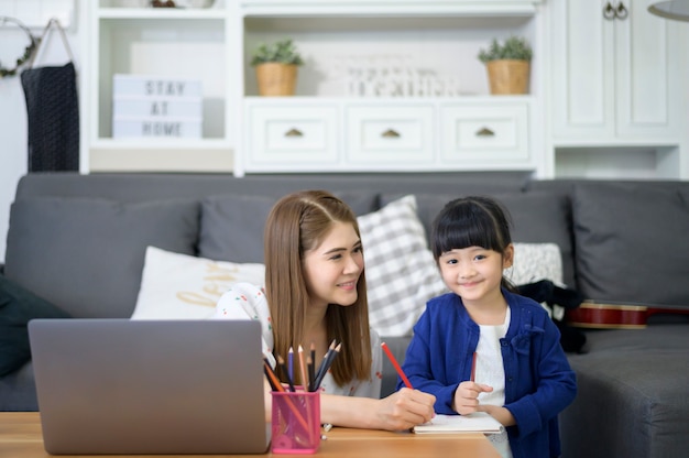 La mamma e la figlia felici asiatiche stanno usando il laptop per studiare in linea via Internet a casa. Concetto di e-learning durante il periodo di quarantena.