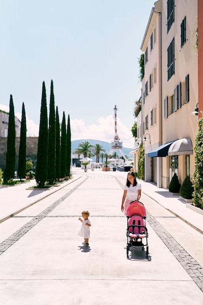 La mamma fa rotolare il passeggino per le strade della città e la bambina cammina nelle vicinanze