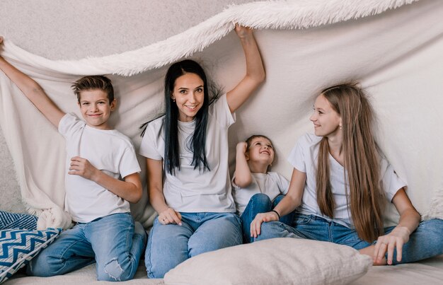 La mamma felice ei suoi bambini piccoli sono seduti sul divano con una coperta bianca sopra la testa, ridendo allegramente. buon fine settimana a casa. gioie familiari