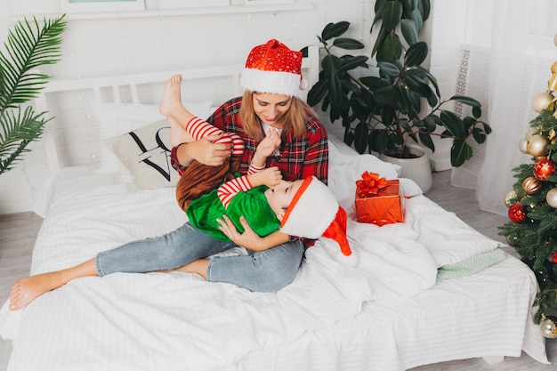La mamma gioca con suo figlio sul letto di casa vicino all'albero di natale. donna e bambino in cappelli di babbo natale. Natale.