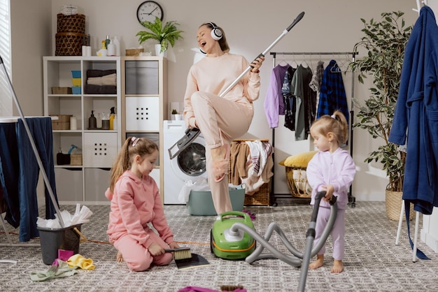 La mamma guarda le figlie che puliscono con le ragazze nel bagno della lavanderia La donna suona il mocio come la chitarra ascolta la musica con le cuffie canta il concerto mentre pulisce