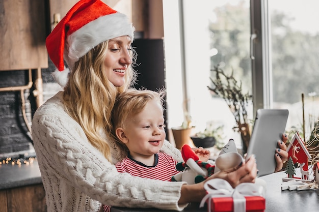 La mamma in un cappello di Babbo Natale festivo di Capodanno con un bambino piccolo si siede a un tavolo con un tablet