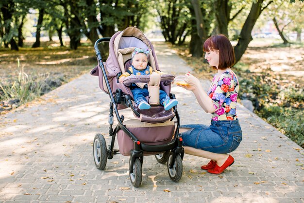 La mamma nutre con il cucchiaio il bambino che cammina con un passeggino nel parco.