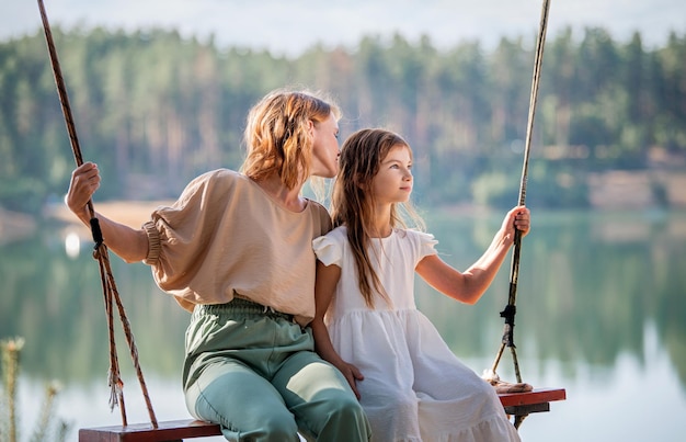 La mamma oscilla con sua figlia su un'altalena di corda sullo sfondo di un lago forestale che riflette il cielo Concetto di famiglia