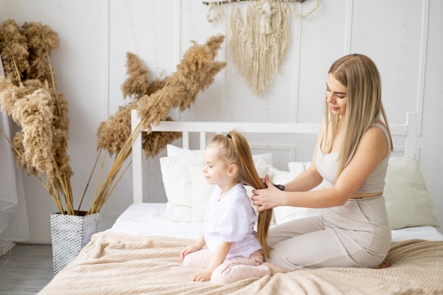 La mamma pettina i capelli di sua figlia a casa sul letto stile di vita tenera relazione di una giovane madre e bambino famiglia felice e maternità che si prendono cura di capelli sani