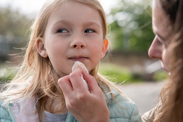 La mamma pulisce la bocca macchiata di una bambina bionda con una giacca nel parco