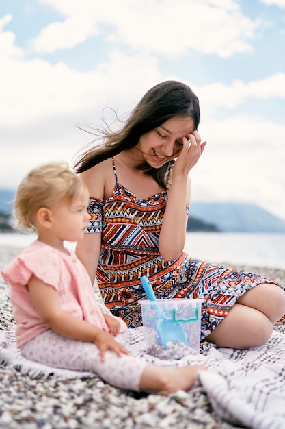 La mamma si raddrizza i capelli al vento mentre è seduta con una bambina su un copriletto sulla spiaggia