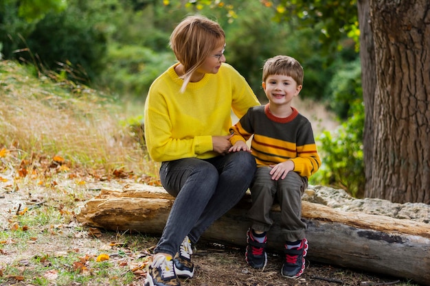 La mamma si siede con suo figlio in maglioni gialli e arancioni seduti fuori nella foresta autunnale